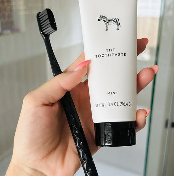 Hand holding a tube of toothpaste labeled 'The Toothpaste' and a black toothbrush in a bathroom setting.